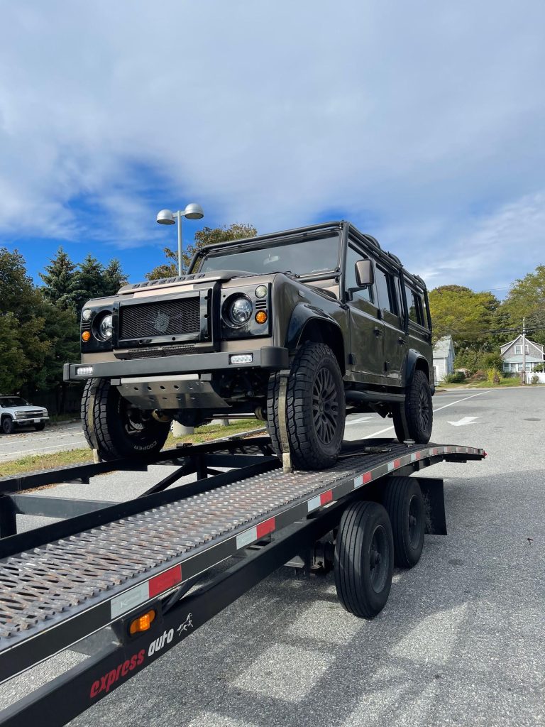 Modified Land Rover Defender 110 on an open auto transport trailer with off-road tires and custom grille